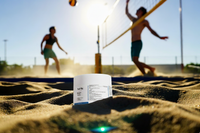 Sol Rx Post-Sun Balm on sand with beach volleyball game in background